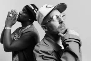Black-and-white portrait of music producers Hit-Boy and The Alchemist. Hit-Boy stands on the left wearing a cap and jewelry, hands clasped in a prayer-like gesture, while The Alchemist faces right in glasses and a cap, resting his chin on his hand in thought. The image captures their creative chemistry and contemplative mood ahead of their 2025 collaboration Goldfish.