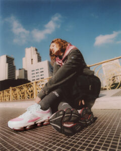 A person sits outdoors on a tiled bridge wearing the New Balance ABZORB 2000 sneakers in the “Rose Sugar with Monarch Burgundy” colourway. The shoes’ pink and white upper contrasts with the city skyline and blue sky in the background, highlighting the sneaker’s bold design and modern cushioning sole.
