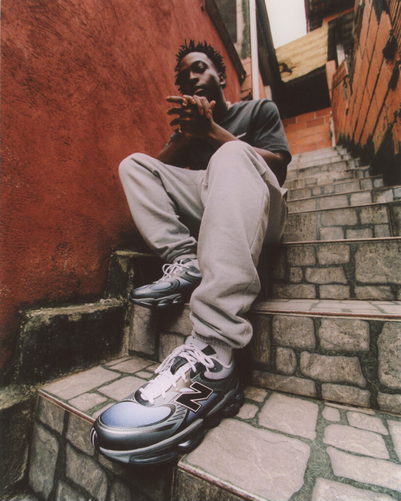 A person sits on stone steps wearing the New Balance ABZORB 2000 sneakers in the “Pearl Grey with Black” colourway. The metallic grey and black shoes stand out against the rustic red and brick alley backdrop, highlighting their sleek, performance-inspired design.