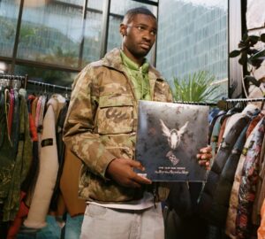 Dave wearing a camouflage Stone Island jacket and light jeans holds a vinyl record titled The Boy Who Played The Harp inside a studio-like space lined with racks of colorful Stone Island outerwear. Sunlight filters through large industrial windows, casting a soft glow that highlights the textures of the garments and the reflective sheen of the record cover.