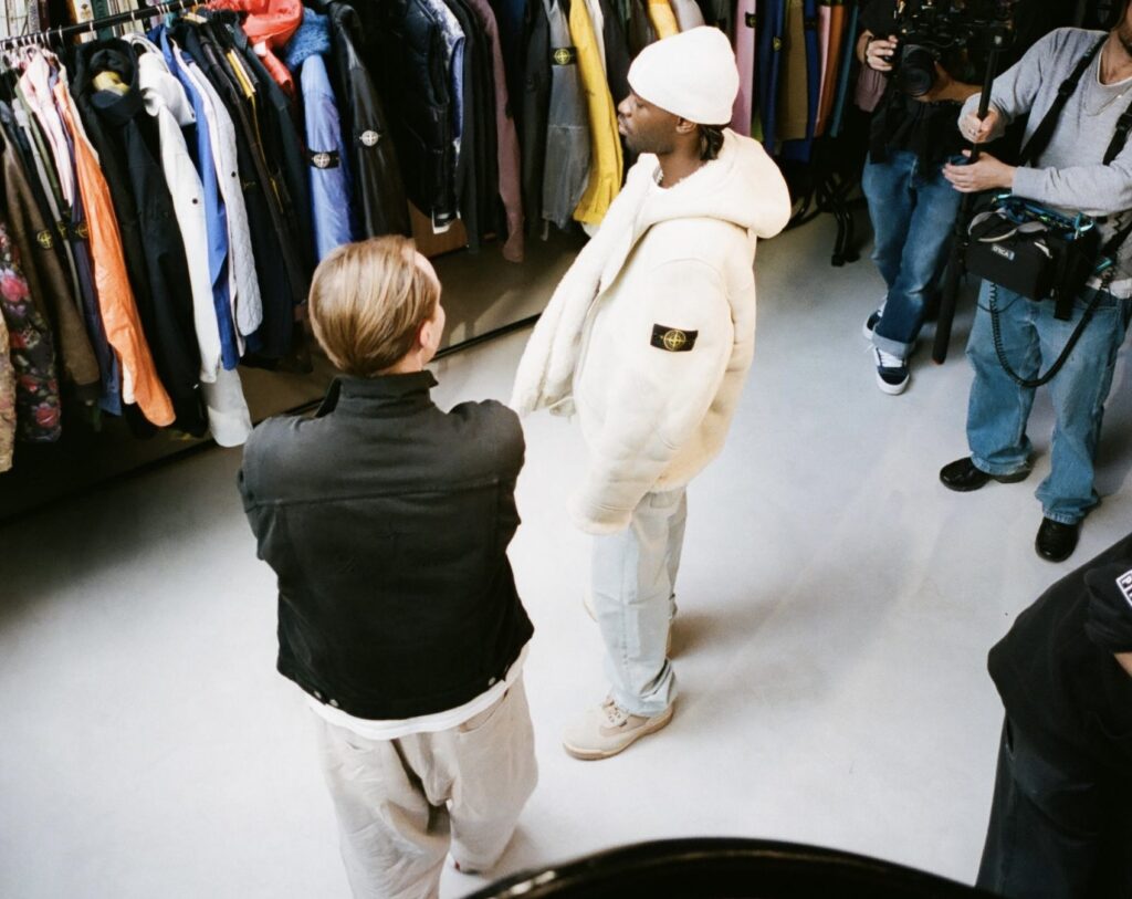 A candid overhead view of two people in conversation inside a studio or showroom, surrounded by racks of colorful Stone Island outerwear. One person wears a white shearling Stone Island jacket and a beanie, while the other, dressed in black, faces them with arms crossed. Nearby, a small camera crew films the interaction, capturing the behind-the-scenes atmosphere of the space.