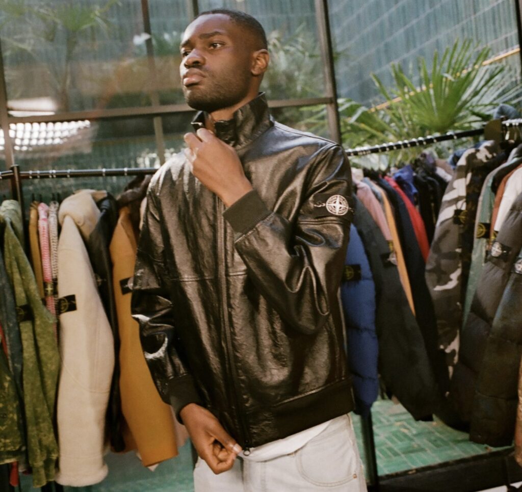 A man wearing a black leather Stone Island jacket stands in front of a clothing rack filled with colorful outerwear, including camouflage, puffer, and shearling pieces. He adjusts the collar of his jacket while looking ahead with a composed expression, framed by natural light streaming through large glass windows and surrounding greenery.