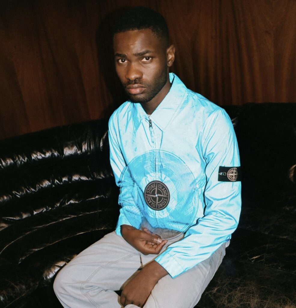 A man sits on a dark leather sofa wearing a light blue Stone Island jacket featuring the brand’s signature compass patch on the sleeve. The glossy texture of the jacket catches the light, contrasting with the deep brown wood-paneled background, creating a moody, introspective atmosphere.
