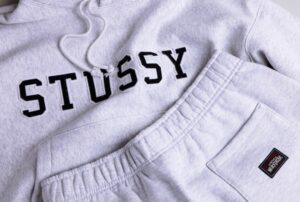 Close-up image of a light grey Stüssy hoodie and sweatpants set, showing the brand’s black varsity-style logo stitched across the chest and a small woven “Authentic Workgear” label on the pocket, highlighting the soft fleece texture and relaxed streetwear aesthetic.
