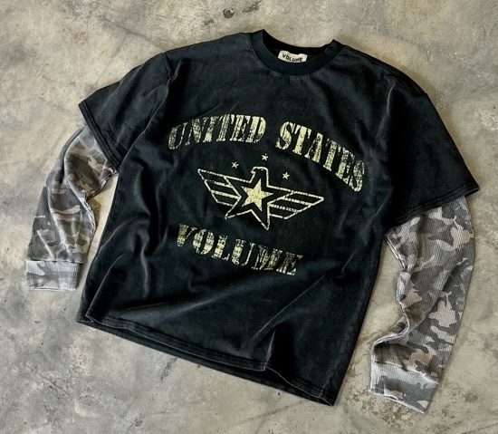 A black double-layer T-shirt featuring distressed stencil text reading “UNITED STATES VOLUME” with a star-and-wings military emblem on the chest, paired with gray camouflage long sleeves, displayed on a concrete background for a rugged streetwear aesthetic.
