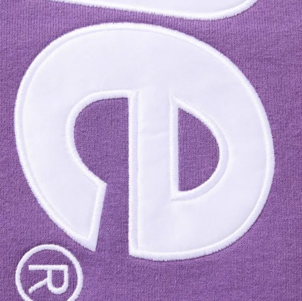 Close-up detail of the Supreme Satin Appliqué Hoodie in purple, showing the textured white satin-stitched lettering on heavyweight cotton fabric. The embroidery highlights the premium craftsmanship and raised logo finish characteristic of Supreme’s FW25 collection.