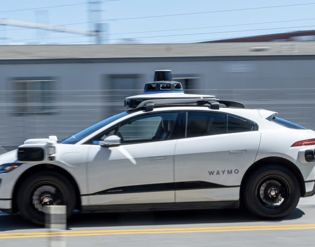 A white Waymo autonomous electric vehicle drives along a city street, equipped with roof-mounted lidar sensors and side-mounted cameras for 360-degree vision—capturing the essence of next-generation robotaxi technology ahead of the company’s 2026 London launch.