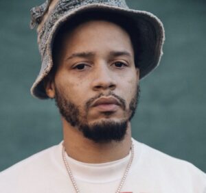 Portrait Veeze wearing a textured denim bucket hat and white shirt, standing against a muted green backdrop. His neutral expression