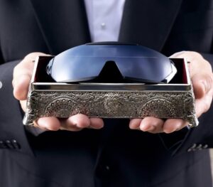 A person in a suit holds an ornate silver box containing a pair of futuristic wrap-around sunglasses with mirrored blue lenses, symbolizing the blend of luxury craftsmanship and modern design.
