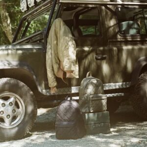 Two Herschel Supply Co Quilted Capsule backpacks in black and olive green rest beside an off-road vehicle with a beige quilted jacket draped over the door, set in a sun-dappled outdoor environment that evokes rugged utility and timeless craftsmanship.