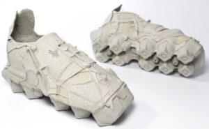 Close-up of Vadim Kibardin’s Egg Box Sneakers, conceptual footwear crafted entirely from recycled molded cardboard. The shoes feature sculptural forms that mimic the contours of egg cartons, with visible seams, layered textures, and geometric recesses emphasizing their raw, paper-based construction. Set against a clean white background, the image highlights the contrast between fragility and structure — a study in sustainable, creative reuse.