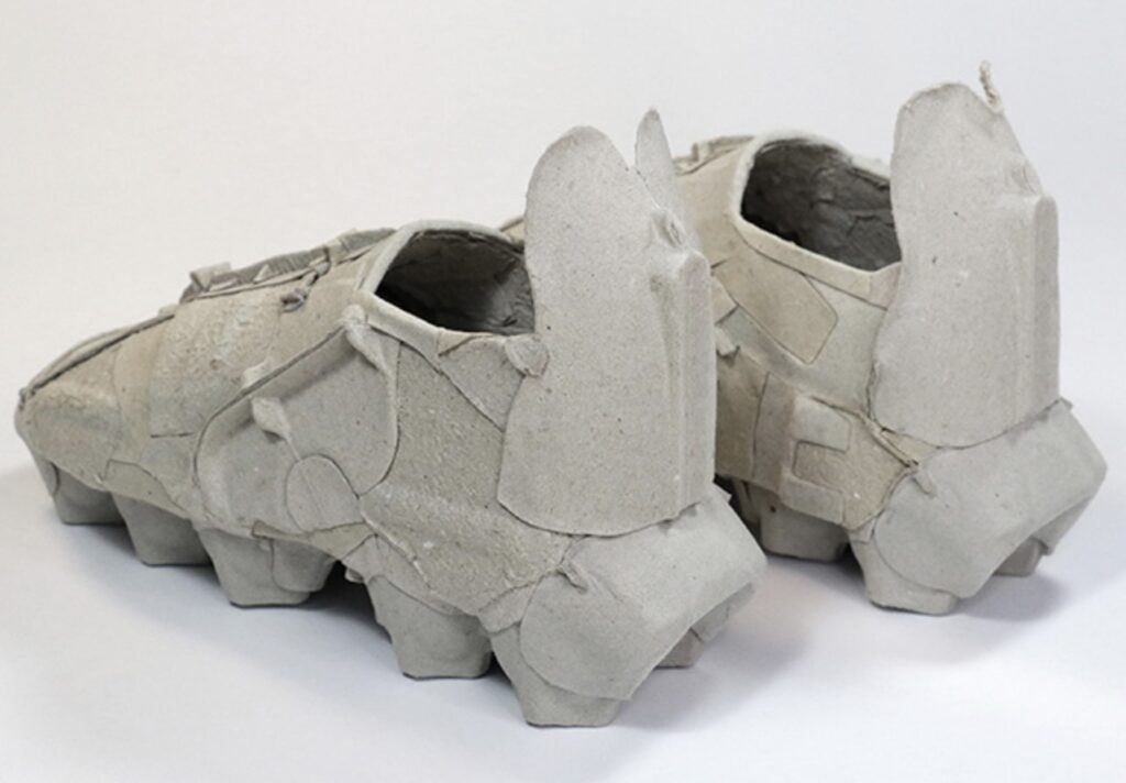 Rear view of Vadim Kibardin’s Egg Box Sneakers, made from molded recycled cardboard. The shoes feature angular, overlapping panels that resemble fragments of egg cartons, with raised heel structures and textured seams emphasizing their handmade construction. Displayed on a neutral white background, the image highlights the sculptural quality and experimental use of waste material as a design medium.