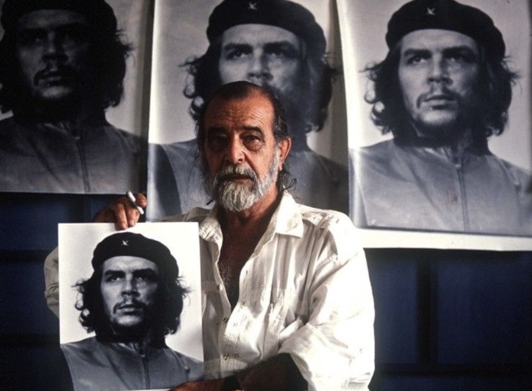 A man with a beard wearing a white shirt holds a black-and-white print of Che Guevara’s famous portrait while standing in front of several larger reproductions of the same image displayed behind him.