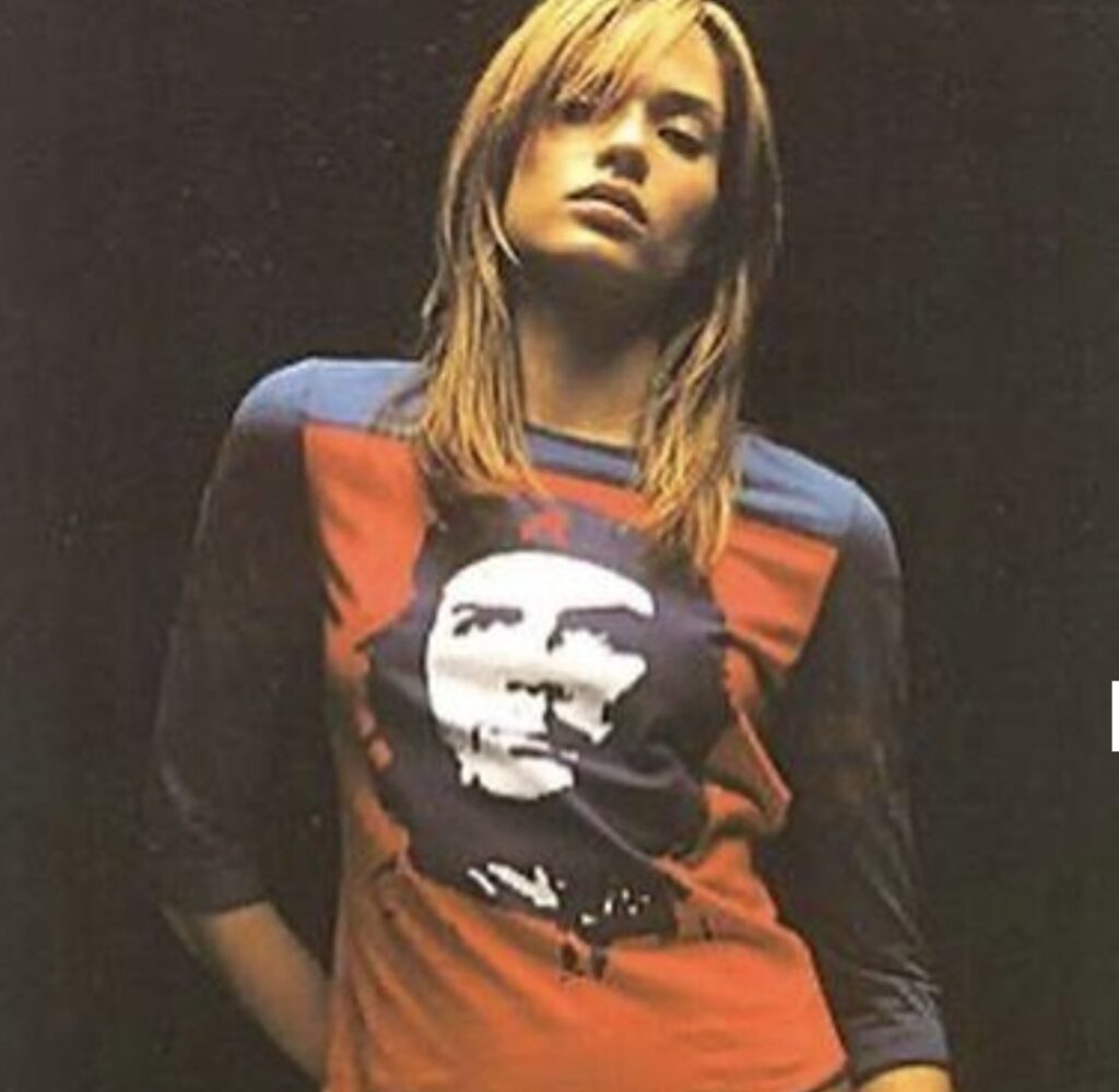 A woman wearing a red and blue long-sleeved shirt featuring a large graphic print of Che Guevara’s face, standing against a dark background with a confident, relaxed pose.