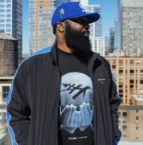 God Shammgod wearing a blue Paper Planes cap and black-and-blue tracksuit jacket from the Shammgod × Planes collection, featuring a black tee printed with a graphic of hands holding a basketball marked by the Paper Planes logo, photographed on a New York rooftop with the city skyline in the background.