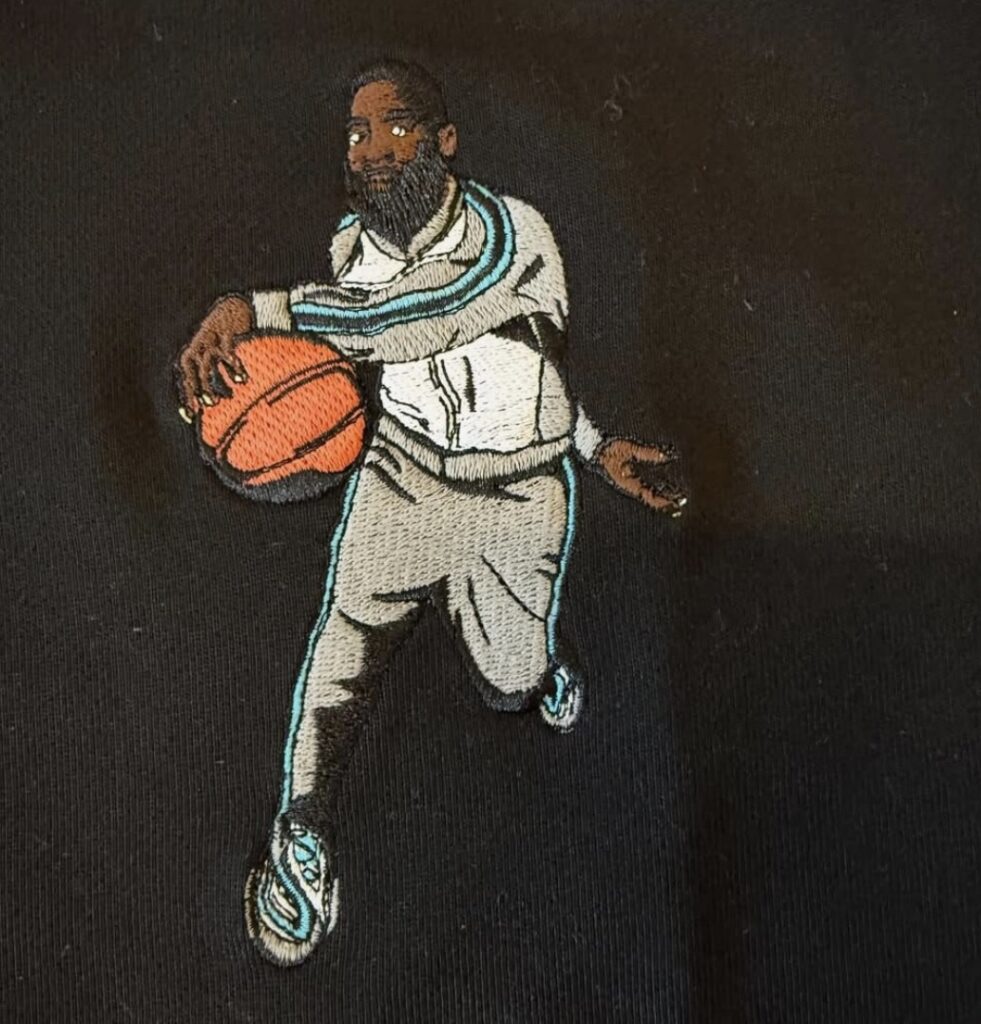 Embroidered patch of a basketball player wearing a grey and teal tracksuit dribbling an orange basketball, depicted on black fabric—part of the Shammgod × Paper Planes collaboration, capturing the signature crossover move in illustrated form.