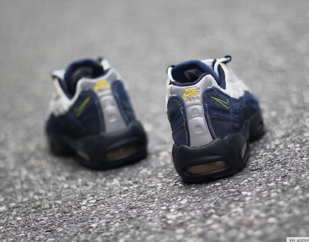 Rear view of the Eric Koston Nike SB Air Max 95 in Obsidian, Speed Yellow, and Summit White, showing the dual Nike SB and Air branding on the reflective heel panel, yellow mini Swoosh embroidery, and visible Air Max cushioning above a dark outsole.