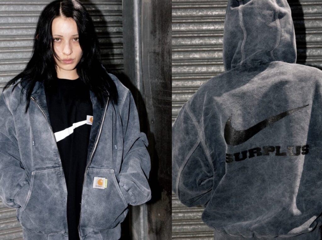 Model wearing the Carhartt WIP × Nike SURPLUS® 2025 washed grey canvas jacket layered over a black tee, showing the front Carhartt label and back view with oversized black Nike Swoosh and ‘SURPLUS’ lettering printed across the hooded jacket.