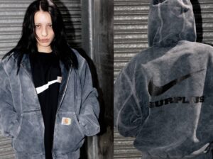 Model wearing the Carhartt WIP × Nike SURPLUS® 2025 washed grey canvas jacket layered over a black tee, showing the front Carhartt label and back view with oversized black Nike Swoosh and ‘SURPLUS’ lettering printed across the hooded jacket.