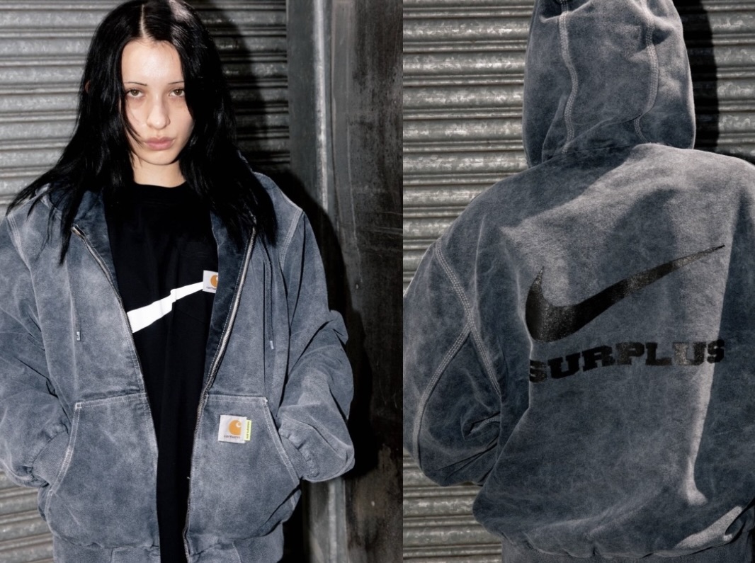 Model wearing the Carhartt WIP × Nike SURPLUS® 2025 washed grey canvas jacket layered over a black tee, showing the front Carhartt label and back view with oversized black Nike Swoosh and ‘SURPLUS’ lettering printed across the hooded jacket.