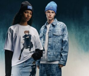 Models wearing pieces from the HUGO × Jujutsu Kaisen capsule collection. The woman on the left wears a white oversized T-shirt printed with an action pose of Nobara Kugisaki layered over a black long-sleeve top, paired with matching light-wash denim featuring the same character motif and a black beanie with the HUGO × Jujutsu Kaisen logo. The man on the right wears a coordinated light-blue denim jacket and pants covered in tonal anime graphics, a white tee, and a bright-blue beanie with embroidered HUGO × Jujutsu Kaisen branding, all set against a dark teal gradient backdrop.