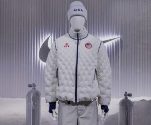 Team USA's official Nike Therma-FIT Air Milano jacket displayed on a mannequin, featuring a quilted inflatable design in white with navy accents, U.S.A. beanie, and Olympic team patch, showcased against a metallic backdrop with the Nike Swoosh logo and air canisters.