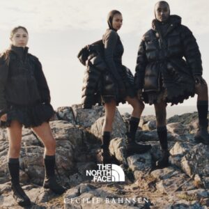 Models wearing The North Face x Cecilie Bahnsen Fall/Winter 2025 collection on a rocky coastal landscape. The group showcases black technical outerwear with sculpted, layered skirts and down-filled jackets, reflecting the collaboration’s fusion of performance design and couture silhouette.