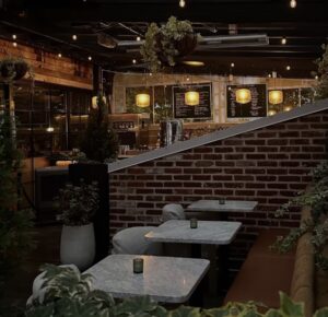 Cozy interior of a boutique cocktail bar featuring marble tables, warm pendant lighting, exposed brick walls, and lush greenery, creating an intimate, modern atmosphere reminiscent of Death & Co’s signature design aesthetic.