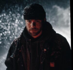 Jake from Publicworksnj wearing a dark jacket and hoodie with a beanie, standing outdoors at night in falling snow. Red light illuminates his face while the snowy background glows softly in the dark.