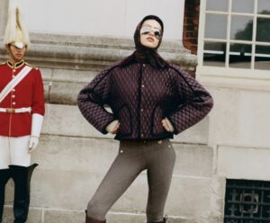 Model wearing a quilted burgundy Burberry jacket with gold buttons, paired with ribbed leggings and leather boots, posing confidently beside a traditional London guard in red uniform and bearskin helmet, set against a historic stone building backdrop.