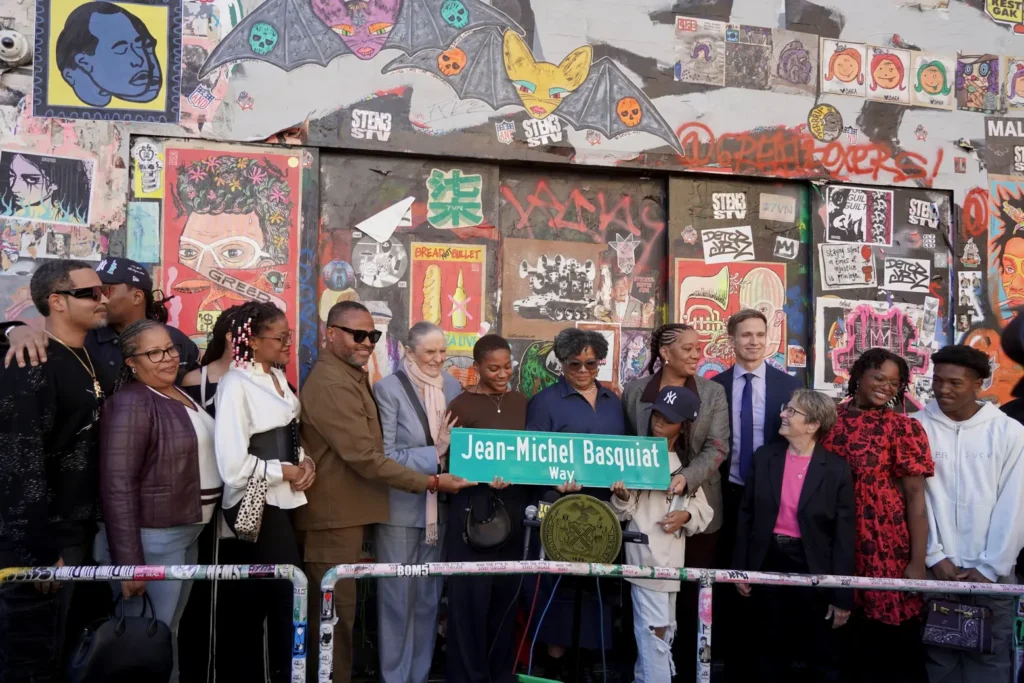 Group photo of the Basquiat family, city officials, and community members gathered in front of Jean-Michel Basquiat’s former studio at 57 Great Jones Street during the unveiling ceremony of “Jean-Michel Basquiat Way.” They hold the new green street sign against a colorful graffiti-covered wall adorned with murals and posters celebrating Basquiat’s artistic legacy.