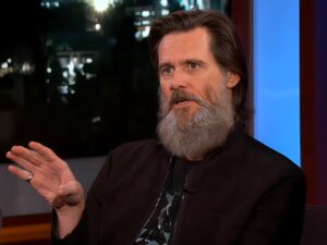 Jim Carrey with a long gray beard, wearing a dark jacket during a television interview, gesturing with his hand while speaking thoughtfully.