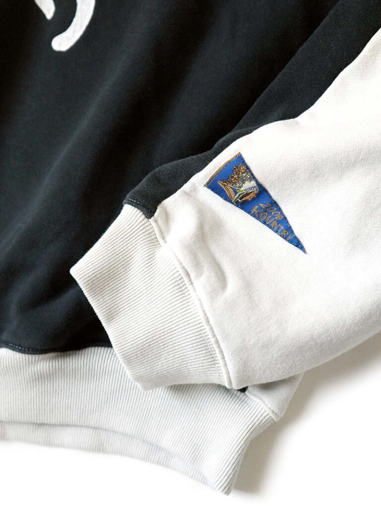Close-up of the KAPITAL Men’s Sweat Knit 2Tones Remake Big Sweatshirt in Ecru and Black, highlighting the ribbed white cuff, contrasting black sleeve panel, and the signature blue KOUNTRY woven label stitched near the wrist.