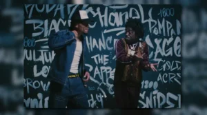 Kodak Black and Chance the Rapper perform side by side in front of a graffiti wall featuring the words “Chanel,” “Chance the Rapper,” and “Pompano.” Kodak wears a vintage-style brown tracksuit and layered jewelry, while Chance sports a denim jacket, white tee, and trucker hat — capturing a blend of street luxury and artistic self-expression that mirrors the theme of Still Get Chanel.