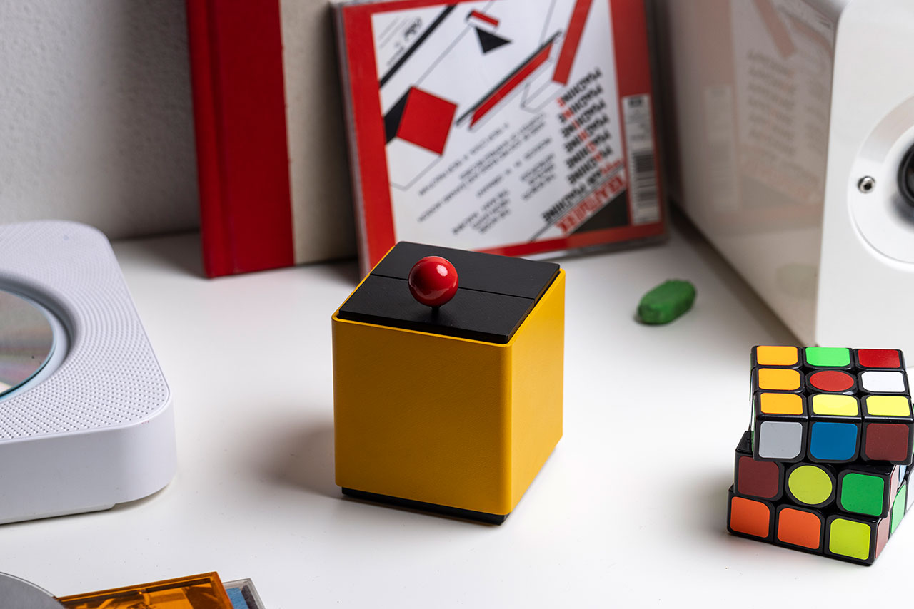 La Machine by Olivier Mével and Elium Studio — a minimalist cube-shaped device in bright yellow with a black top and a glossy red switch. The object sits on a white desk beside a Rubik’s Cube, books, and a speaker, blending playful postmodern design with everyday surroundings.