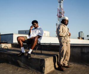 Mick Jenkins and EMIL pose on a rooftop under a clear sky, one seated in a white shirt and shorts with sunglasses, the other standing in a beige hoodie and pants with a cap, urban skyline and radio tower in the background.