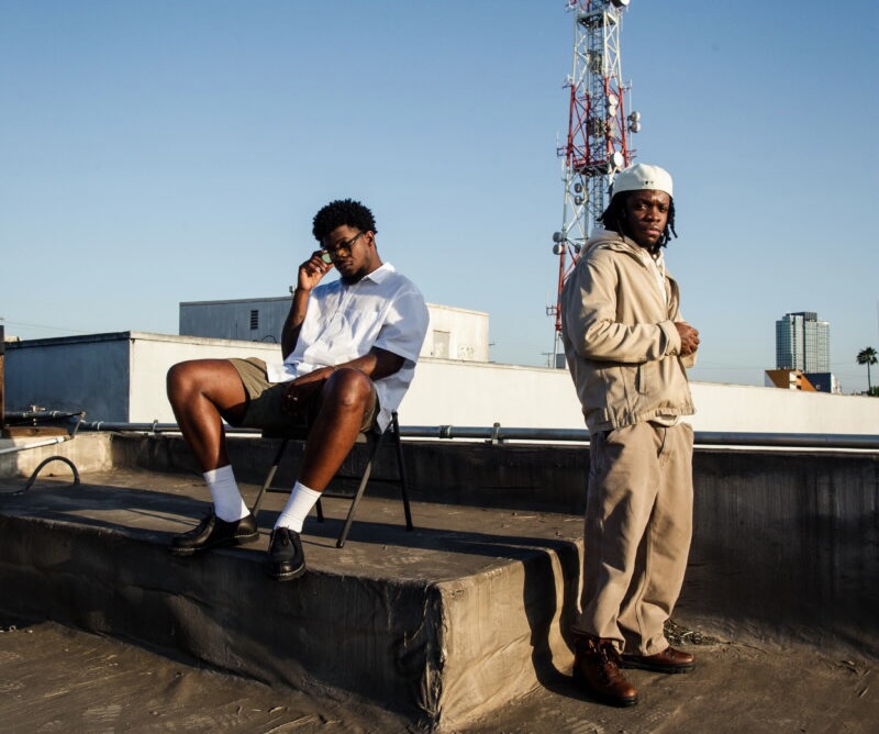 Mick Jenkins and EMIL pose on a rooftop under a clear sky, one seated in a white shirt and shorts with sunglasses, the other standing in a beige hoodie and pants with a cap, urban skyline and radio tower in the background.