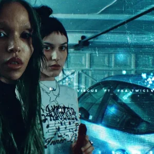 In the blue haze of a garage, Oklou and FKA twigs face the camera, their expressions calm and unreadable. Metallic light glances off a parked car behind them, soft reflections tracing the edges of shadow. The air feels still, charged with the quiet tension of their connection “viscus.”