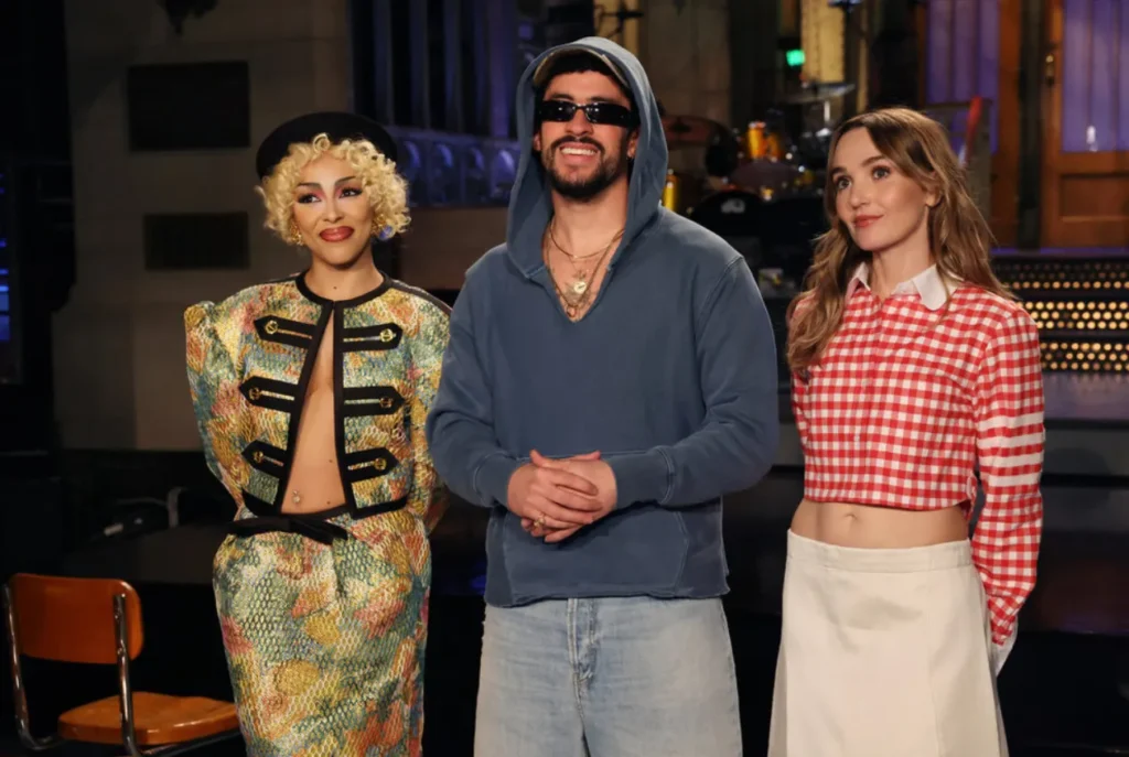 Bad Bunny smiles on stage during Saturday Night Live rehearsals, standing between two cast members—one in a colorful patterned jacket and beret, the other in a red gingham top and white skirt—during the lead-up to his 2026 Super Bowl halftime show monologue.