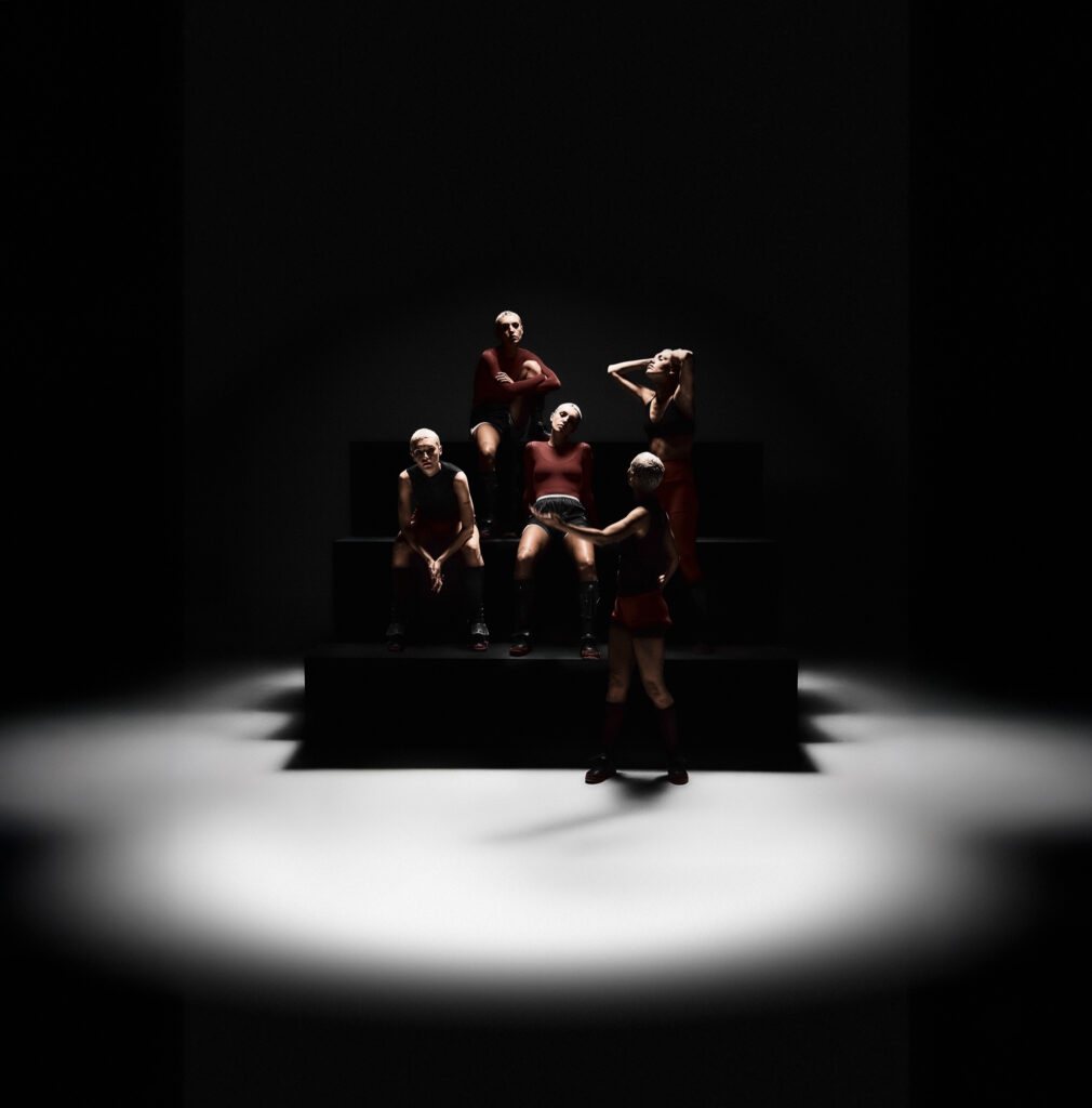 Editorial campaign image for Saucony SILO Fall/Winter 2025, featuring five models with short platinum hair styled in athletic-inspired red and black outfits, positioned dramatically on tiered black platforms under a spotlight in a minimal dark studio setting.