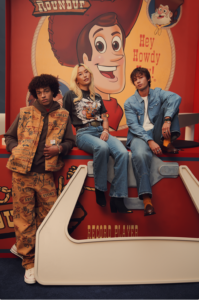Models showcase the Levi’s x Toy Story 30th Anniversary Collection, featuring a quilted vest with all-over Toy Story graphics, a black Woody graphic T-shirt with denim jeans, and a classic Levi’s denim-on-denim look, posed in front of a giant Woody backdrop with the phrase “Hey Howdy Hey.”