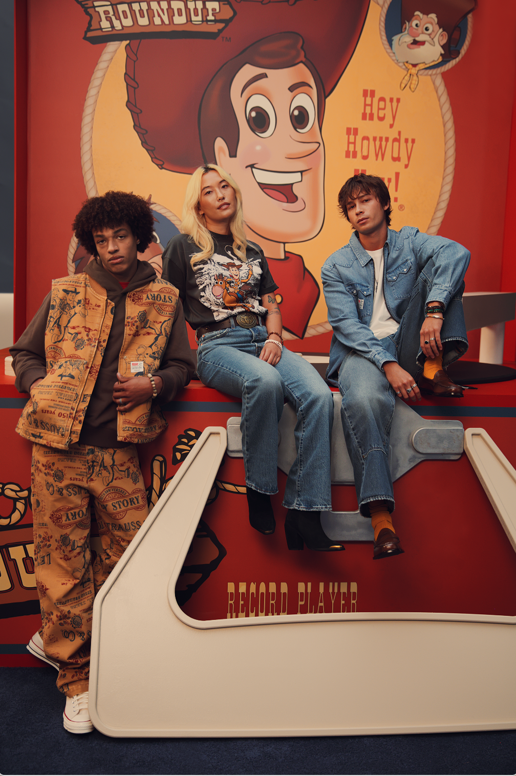 Models showcase the Levi’s x Toy Story 30th Anniversary Collection, featuring a quilted vest with all-over Toy Story graphics, a black Woody graphic T-shirt with denim jeans, and a classic Levi’s denim-on-denim look, posed in front of a giant Woody backdrop with the phrase “Hey Howdy Hey.”