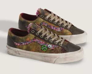 Vans Premium Old Skool “Souvenir” in Warm Brown — a pair of low-top sneakers featuring a distressed brown canvas base with glossy leather overlays, multicolored tweed side stripes, tan laces, and vintage-style souvenir pins including a green Vans star and peace symbol on the lateral side, all set on an aged off-white vulcanized sole.