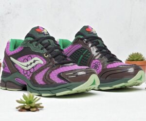 Close-up of the Saucony x Jae Tips ProGrid Triumph 4 shoes in vivid magenta and forest green tones, featuring floral embroidery on the sides, reflective overlays, and mint-green outsoles. The design blends glossy brown accents with textured fabric panels, symbolizing Jae Tips’ expressive, Bronx-inspired creativity and signature Savior Worldwide storytelling. Small succulents decorate the minimalist studio backdrop.