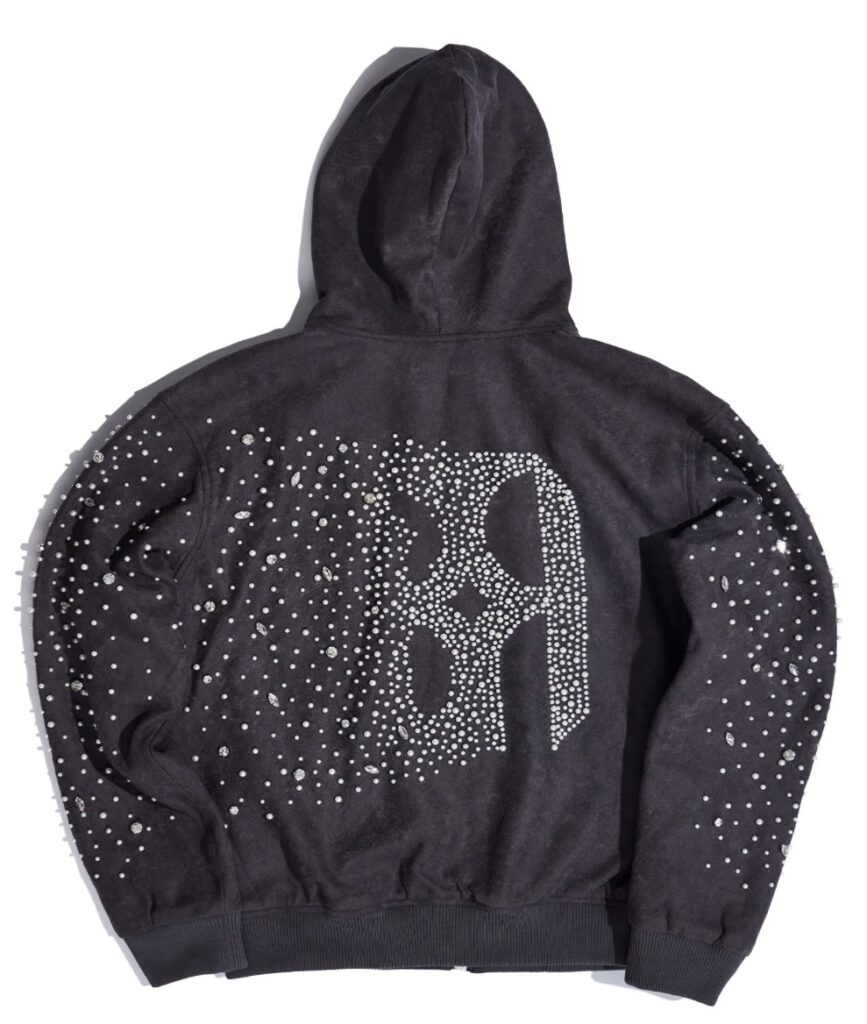 Back view of a charcoal-grey hoodie embellished with pearl and crystal studs forming a large ornate “B” design at the center. The scattered rhinestones across the sleeves and shoulders create a shimmering, galaxy-like effect against the soft suede-textured fabric.