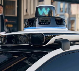 Close-up of a Waymo autonomous vehicle’s advanced sensor array, featuring lidar, radar, and cameras atop the roof with the illuminated “W” logo—showcasing the precision engineering behind Waymo’s AI-driven robotaxi technology ahead of its 2026 London debut.