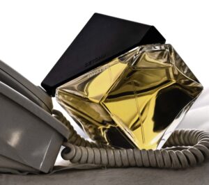 A sculptural glass perfume bottle filled with golden liquid, labeled “SETCHU,” featuring a sharp black geometric cap. The bottle rests against a coiled vintage telephone cord on a neutral surface, evoking a minimalist yet tactile sense of modern haute.