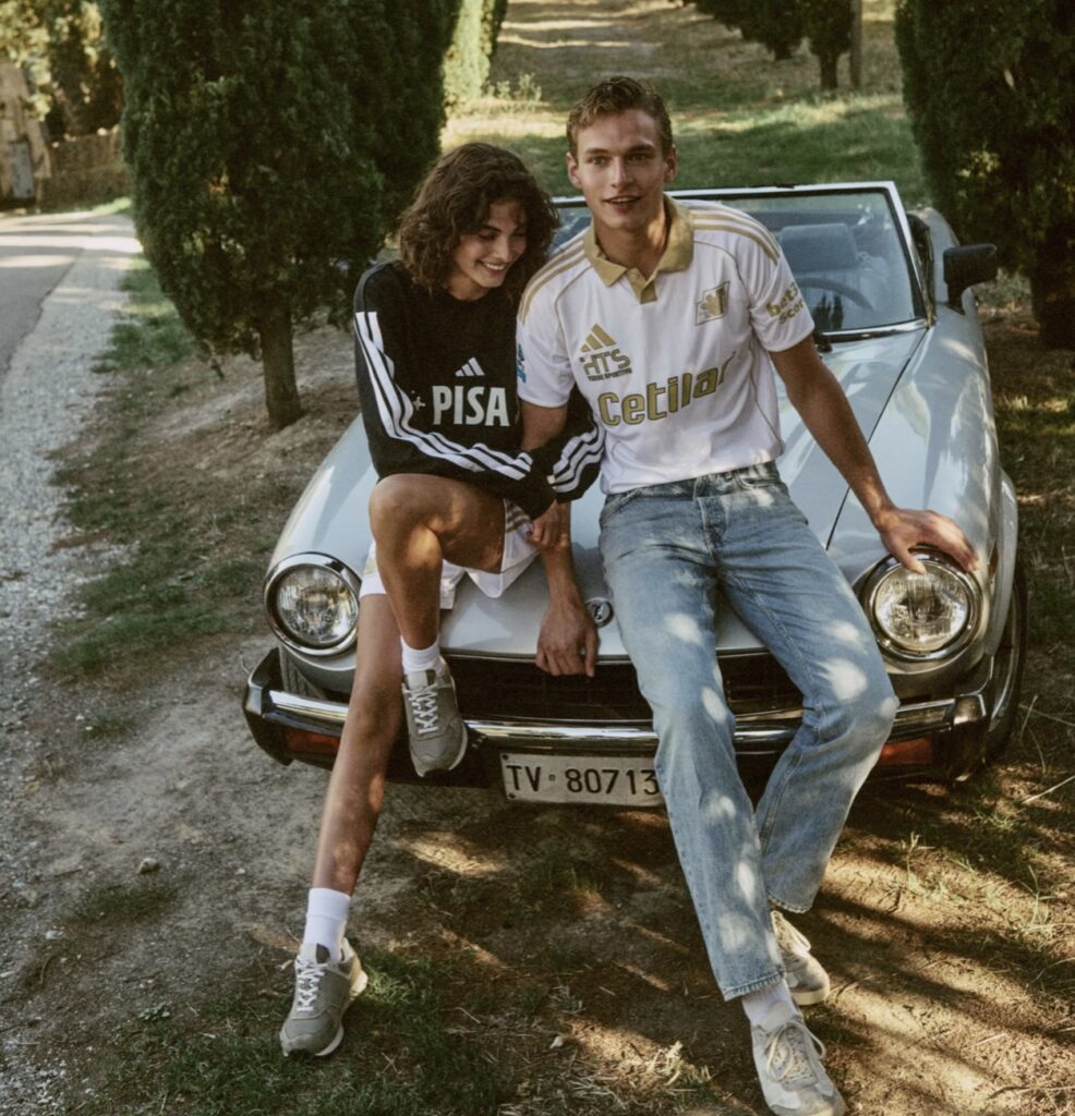 Two models sit on the hood of a vintage convertible wearing Pisa SC x adidas 2025/26 collection; the man sports the white and gold Third Kit jersey with sponsor Cetilar, while the woman wears a black PISA sweatshirt with white adidas stripes, captured on a sunlit Tuscan road lined with cypress trees.