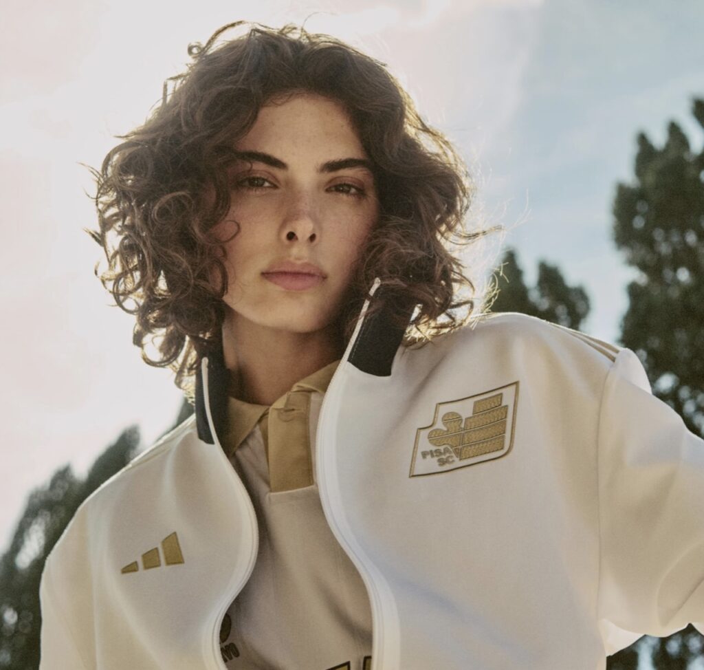 Model wearing the Pisa SC 2025/26 Third Kit by adidas, featuring the revived 1980s crest in gold embroidery on a white zip-up jacket with beige detailing, photographed outdoors under soft sunlight with trees in the background.