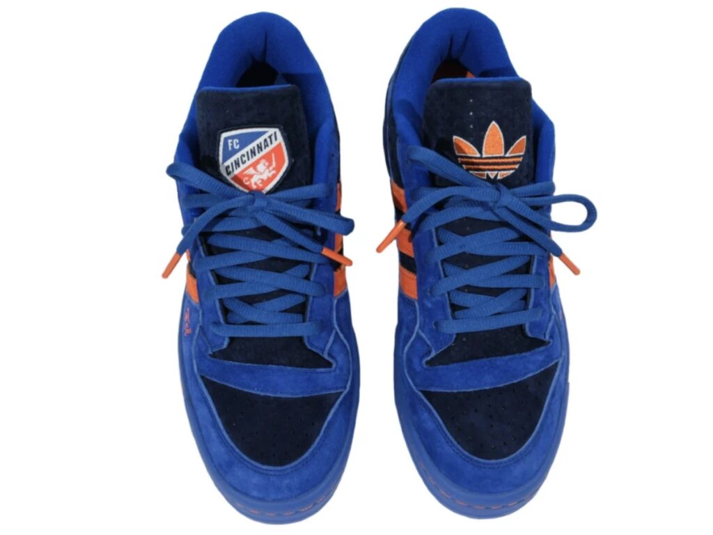 Top-down view of the adidas Forum Low “All For Cincy” Blue Kit edition, showing navy and royal-blue suede uppers, orange adidas Three Stripes, and mismatched tongue logos—one featuring the FC Cincinnati crest and the other the orange adidas Trefoil—finished with blue laces and orange lace tips.